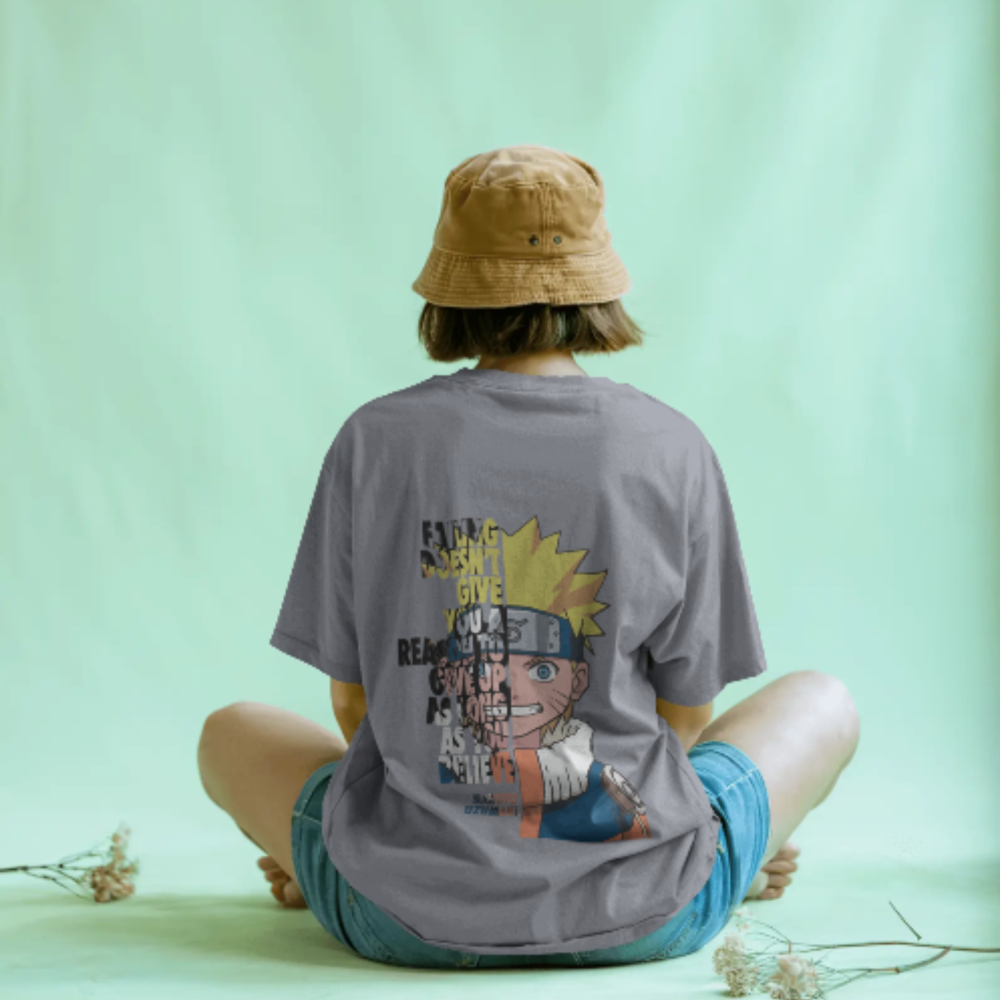 A person sitting cross-legged facing away from the camera, wearing a grey oversized t-shirt with an illustration of Naruto Uzumaki and motivational text. The background is a pastel green and the person wears a tan bucket hat and denim shorts.