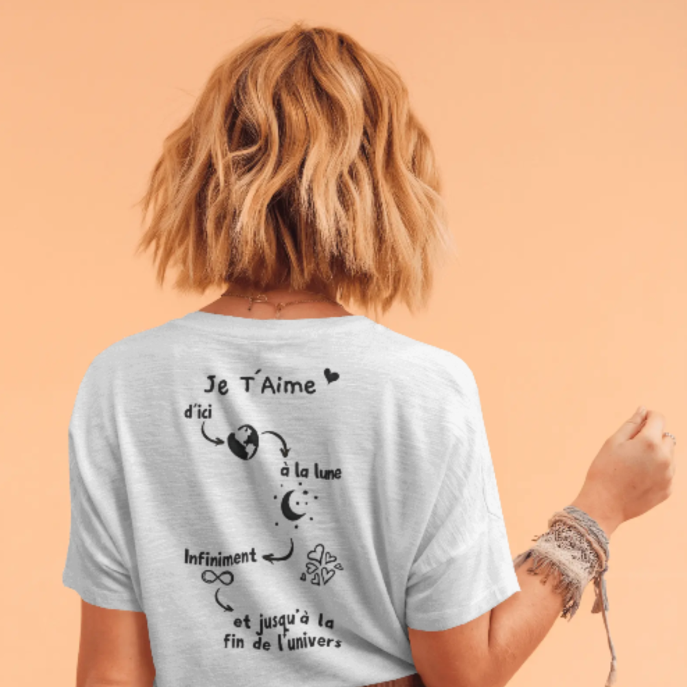 A woman wearing a white t-shirt with a romantic French quote and celestial-themed illustrations on the back, including a moon, Earth, infinity symbol, and stars. The quote reads: "Je T’aime d’ici à la lune, infiniment, et jusqu’à la fin de l’univers.