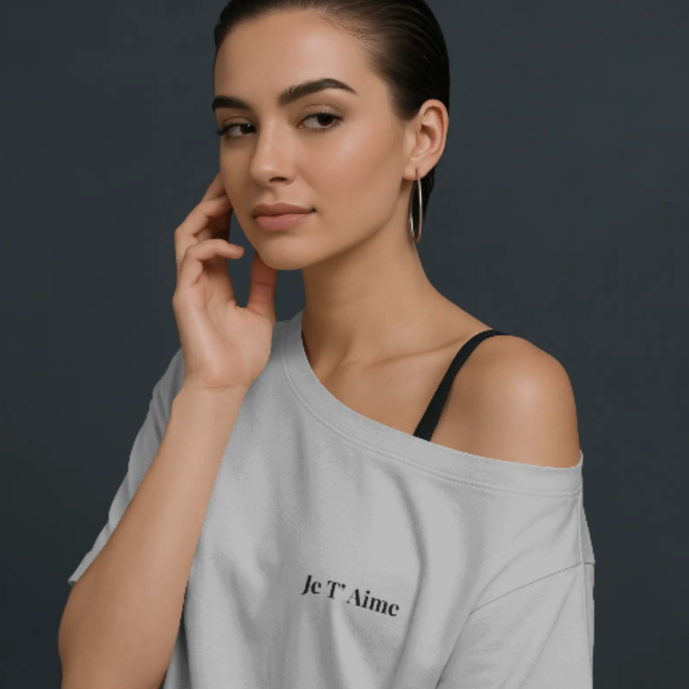 A woman wearing a soft off-shoulder white t-shirt with the French phrase “Je T’aime” printed on the front left chest. She has a confident expression, hoop earrings, and sleek hair against a dark studio background.