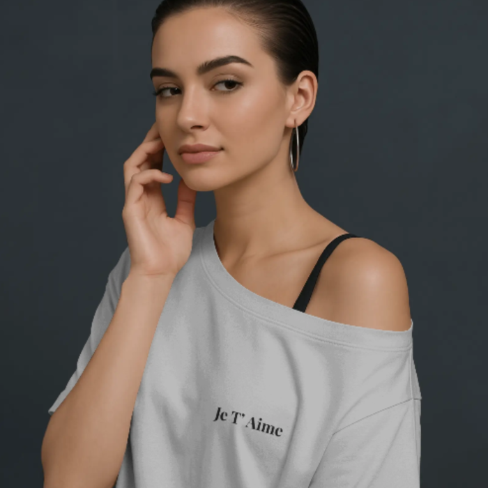 A woman wearing a soft off-shoulder white t-shirt with the French phrase “Je T’aime” printed on the front left chest. She has a confident expression, hoop earrings, and sleek hair against a dark studio background.