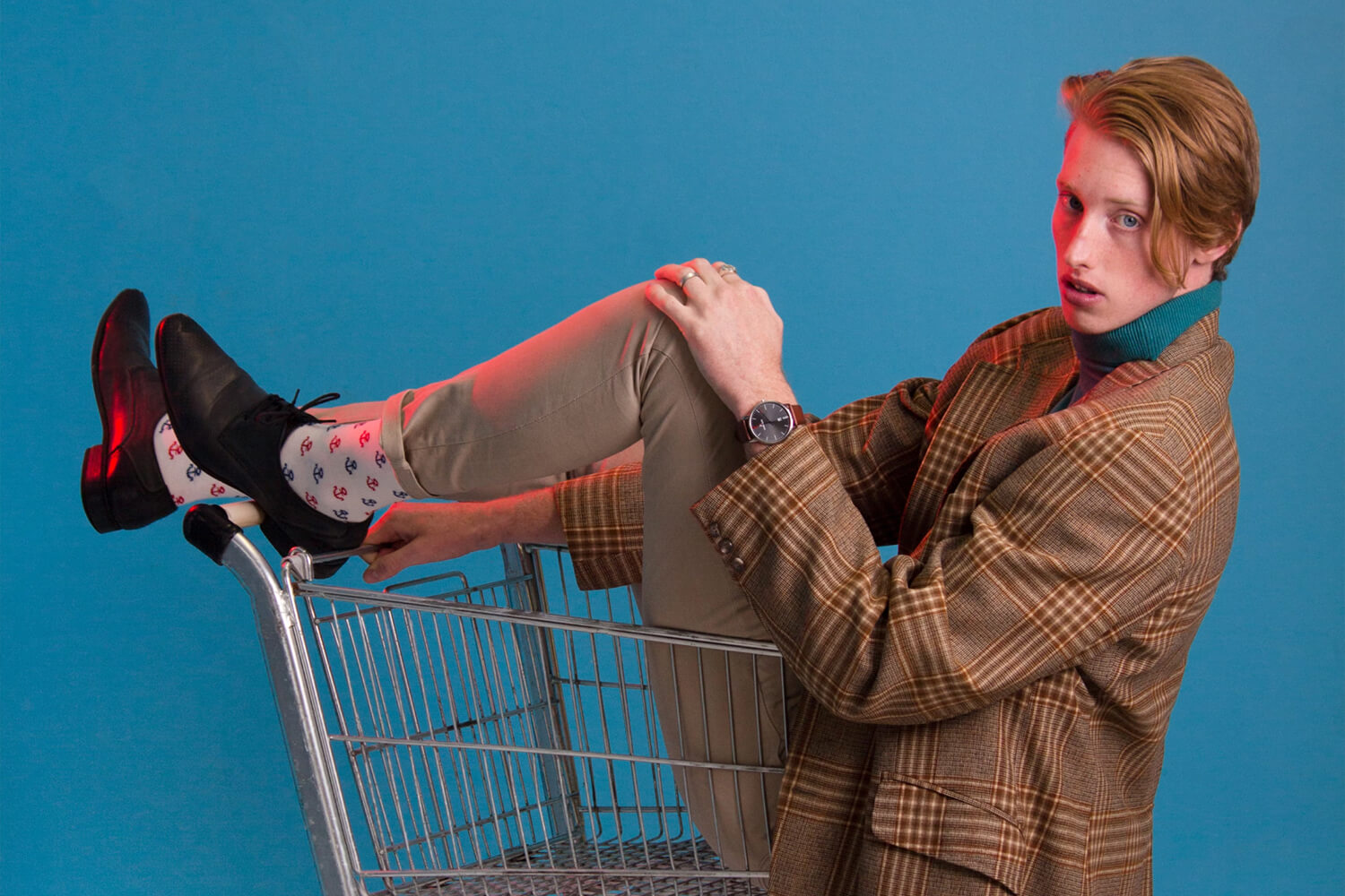 A person sitting in a shopping cart with one leg propped up, wearing a plaid blazer, beige pants, black shoes, and playful patterned socks, my account against a vibrant blue background.