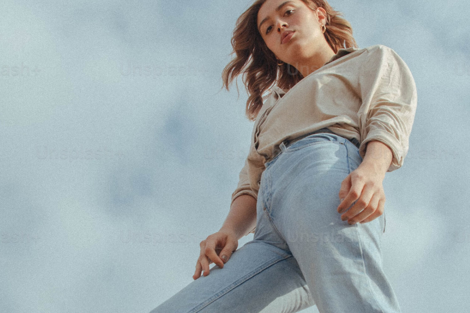 "A person standing confidently, looking down while wearing a beige shirt and light blue jeans, with a cloudy sky in the background.