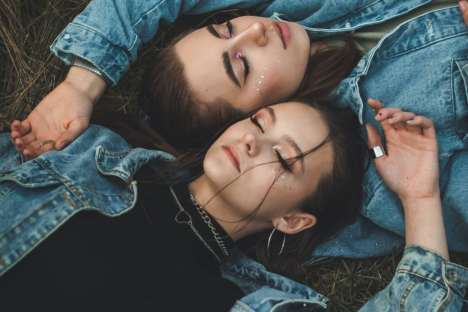 Two young women lying on the grass, wearing denim jackets, with glitter makeup around their eyes, eyes closed, looking peaceful and relaxed.