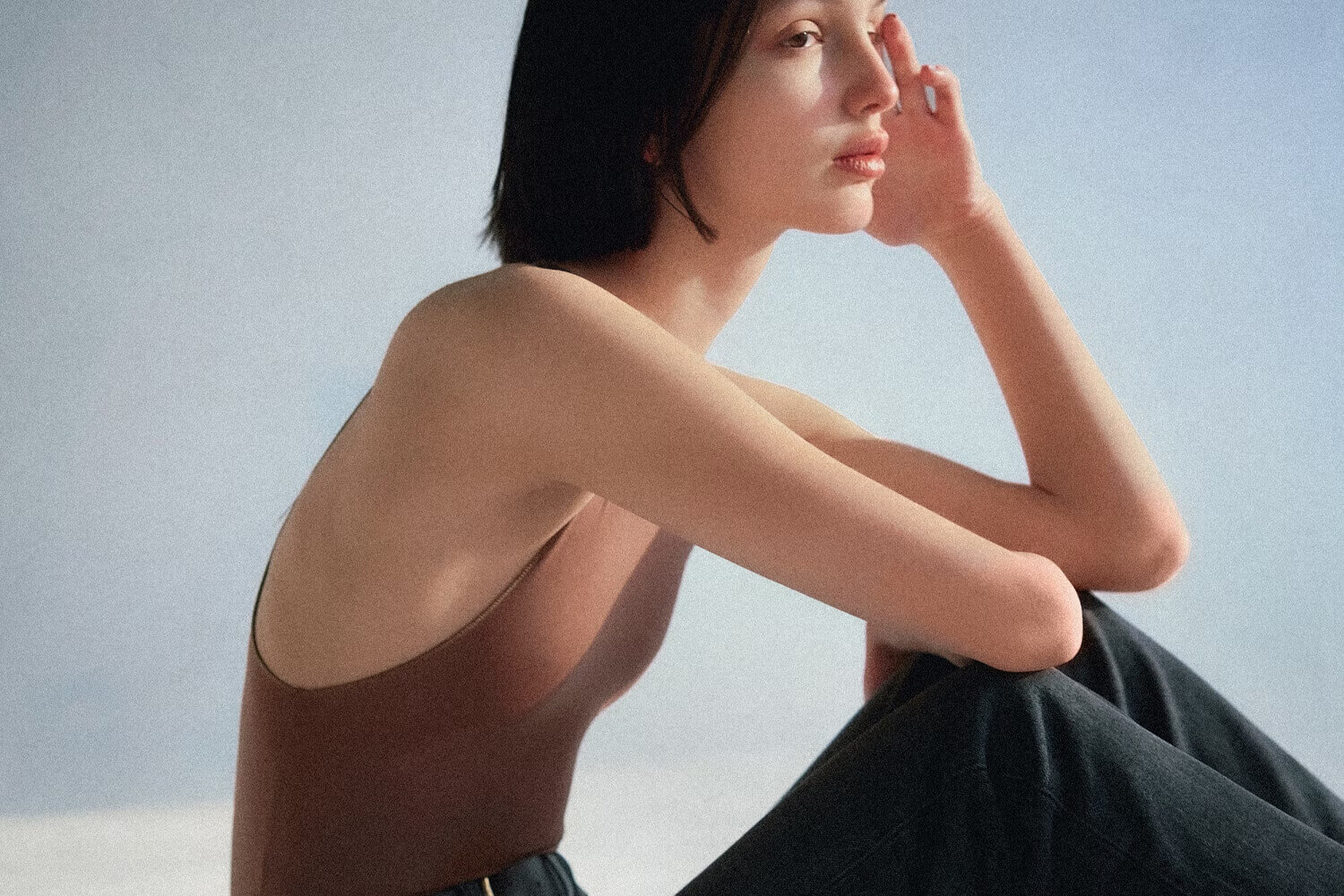 A woman in a brown sleeveless top, sitting with one hand on her chin, gazing thoughtfully against a soft, pastel background.