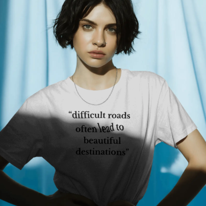 A woman wearing a white T-shirt with the Motivational Quote “Difficult roads often lead to beautiful destinations” in a clean, serif font. She stands in dramatic lighting, adding emphasis to the empowering message.