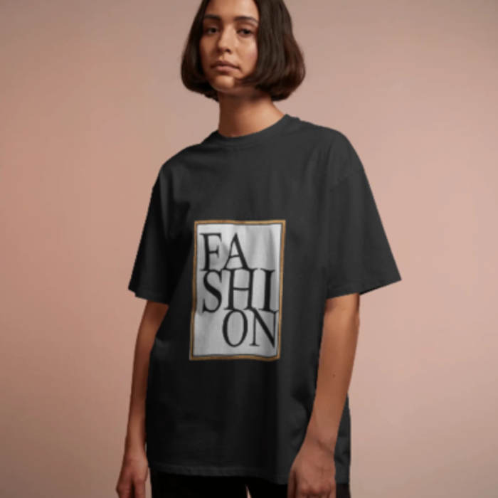A woman with a straight bob haircut wearing a black oversized T-shirt with a large graphic text that reads “Glam Life ” in a tall serif font, arranged vertically within a gold-outlined box. The background is a soft blush pink.