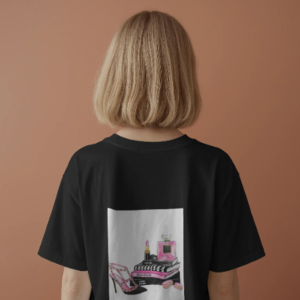 A woman with shoulder-length blonde hair wearing a black T-shirt featuring a graphic on the back of pink high heels, perfume, lipstick, fashion books, and macarons—styled in a chic, girly aesthetic.