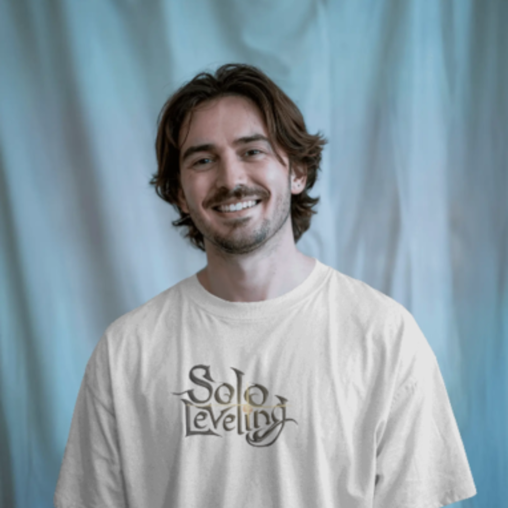 A man wearing a white T-shirt with the text “Solo Leveling” printed on the front in a fantasy-style serif font. He’s smiling confidently against a cool-toned, softly draped background.