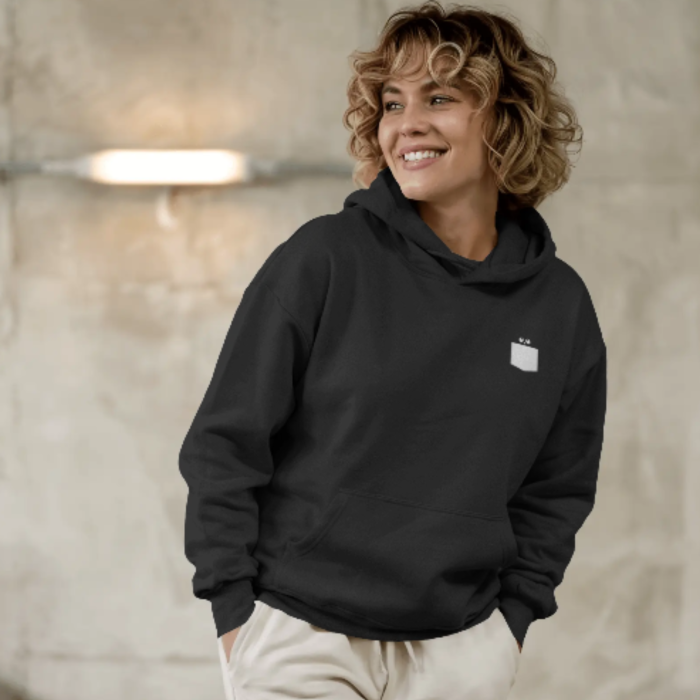 A woman smiling while wearing a black hoodie with a small white pocket-style of Fierce Dog graphic on the left chest area, featuring three tiny stars above a minimalist blank space. The vibe is casual and cozy against a concrete backdrop.