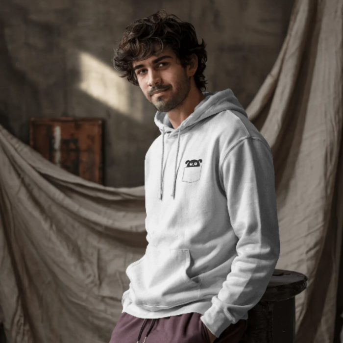 A man wearing a light gray hoodie with a small Puppy Pocket out from a printed front pocket design, posing casually against a soft, industrial-style backdrop.