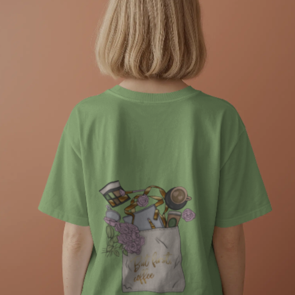 A woman facing away, wearing a green tee with a playful back print showing an overflowing tote bag filled with coffee, books, flowers, ribbon, and sweets. The bag reads, “But first, coffee” in script.