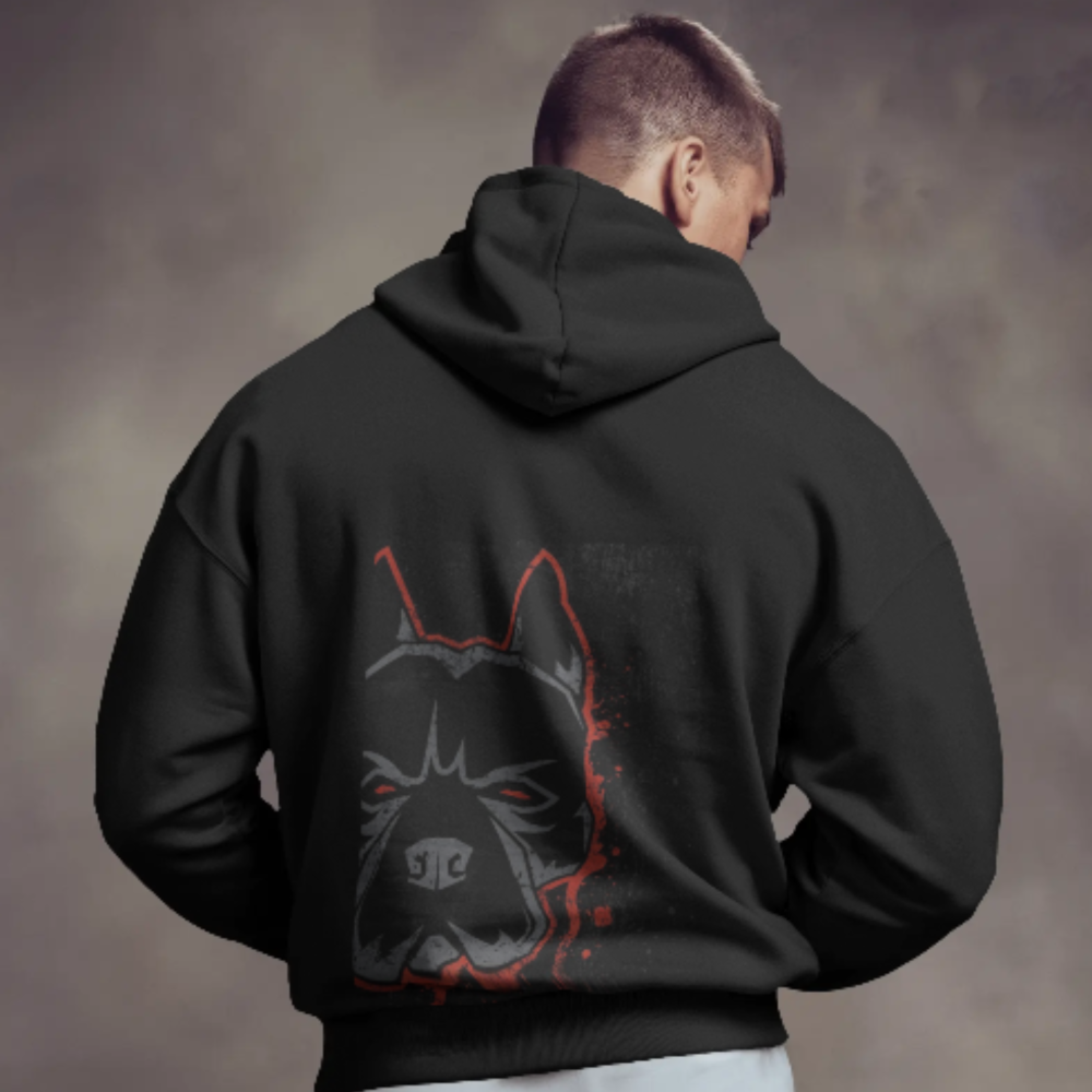 A man wearing a black hoodie with a large back graphic of a fierce, stylized dog’s face in black, red, and grey tones. The intense stare and shadowed detailing give the design an edgy, street-art aesthetic.