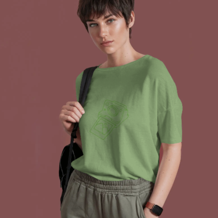 A woman in a light green oversized T-shirt featuring a Coffee simple, monochrome line art graphic of a vintage instant camera on the chest. She’s styled casually with a backpack, giving off chill creative vibes.