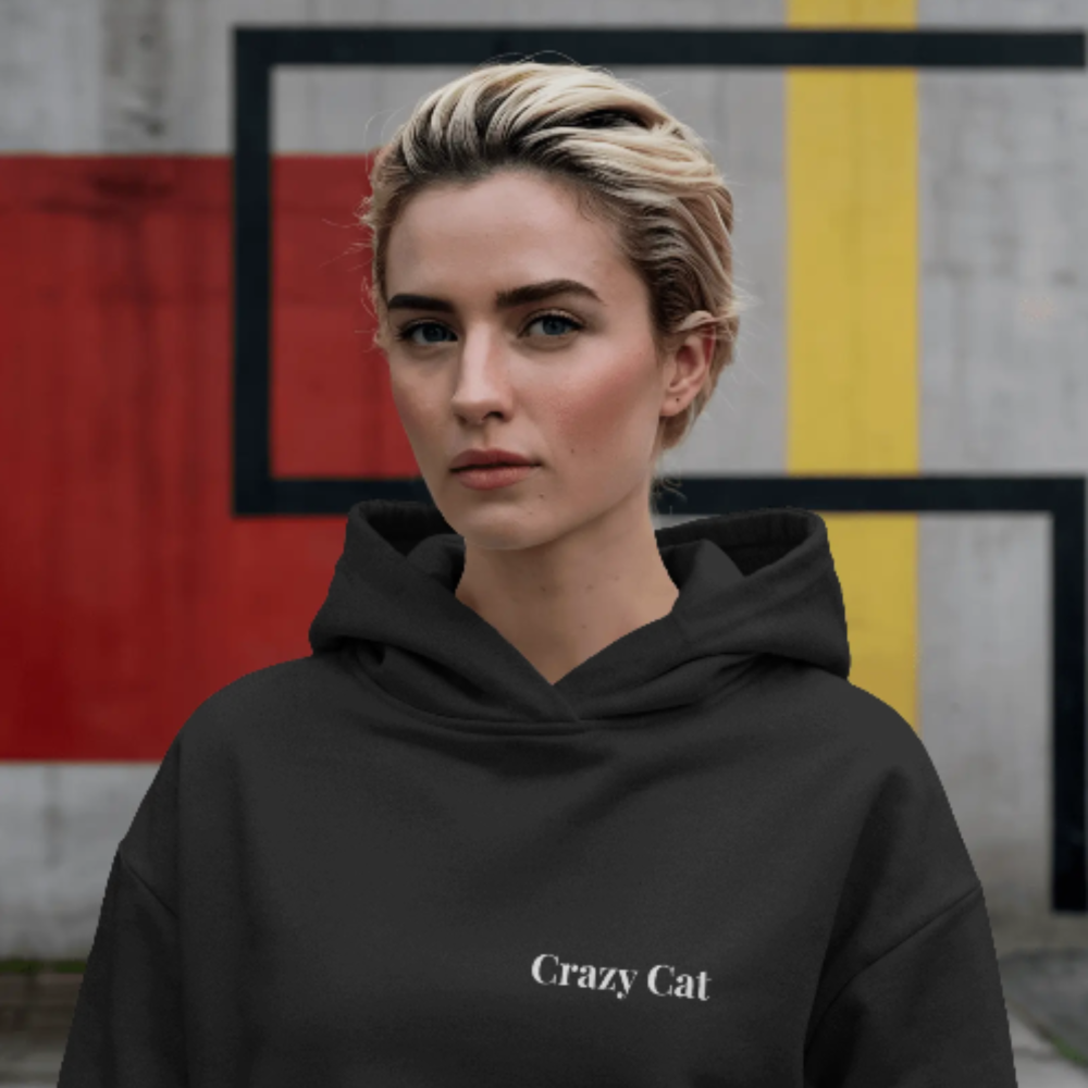 A woman wearing a black hoodie with the clean, minimalist text “Crazy Cat” printed on the front. She’s standing against a modern, geometric wall with confident energy and urban edge.