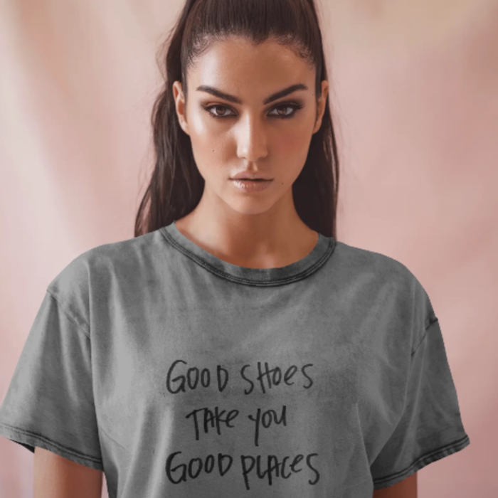 A woman wearing a washed grey T-shirt with the quote “Good shoes take you good places” written in a casual, hand-lettered style. The background is softly pink, adding a warm tone to the minimalist, stylish vibe.