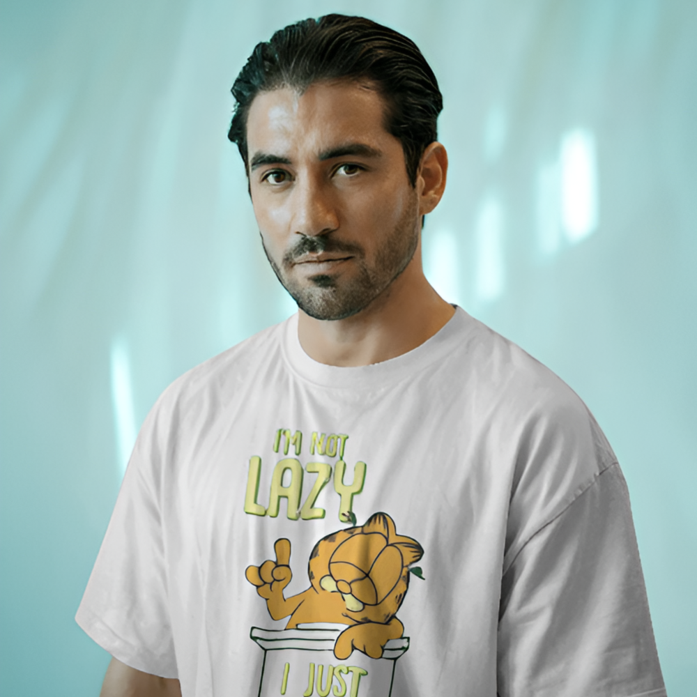 A man wearing a white T-shirt with a cartoon print of Garfield lounging on a ledge and the text "I’m not lazy, I just enjoy doing nothing" in green and yellow. The background has soft teal-blue lighting for a relaxed vibe.