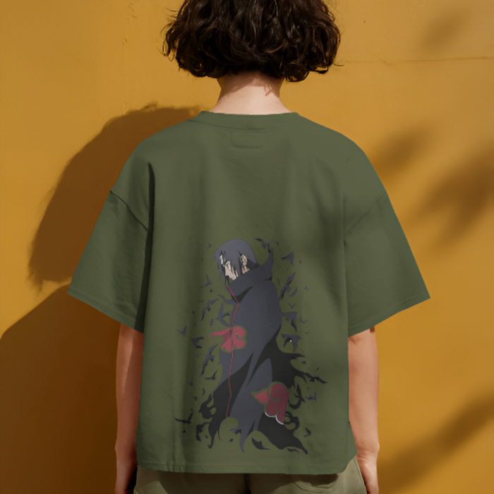 A person wearing an olive green T-shirt with a large back graphic of Itachi Uchiha from Naruto in his Akatsuki cloak, surrounded by dark aura and red cloud symbols. The character is facing sideways in a moody pose, against a mustard yellow wall with natural shadows.