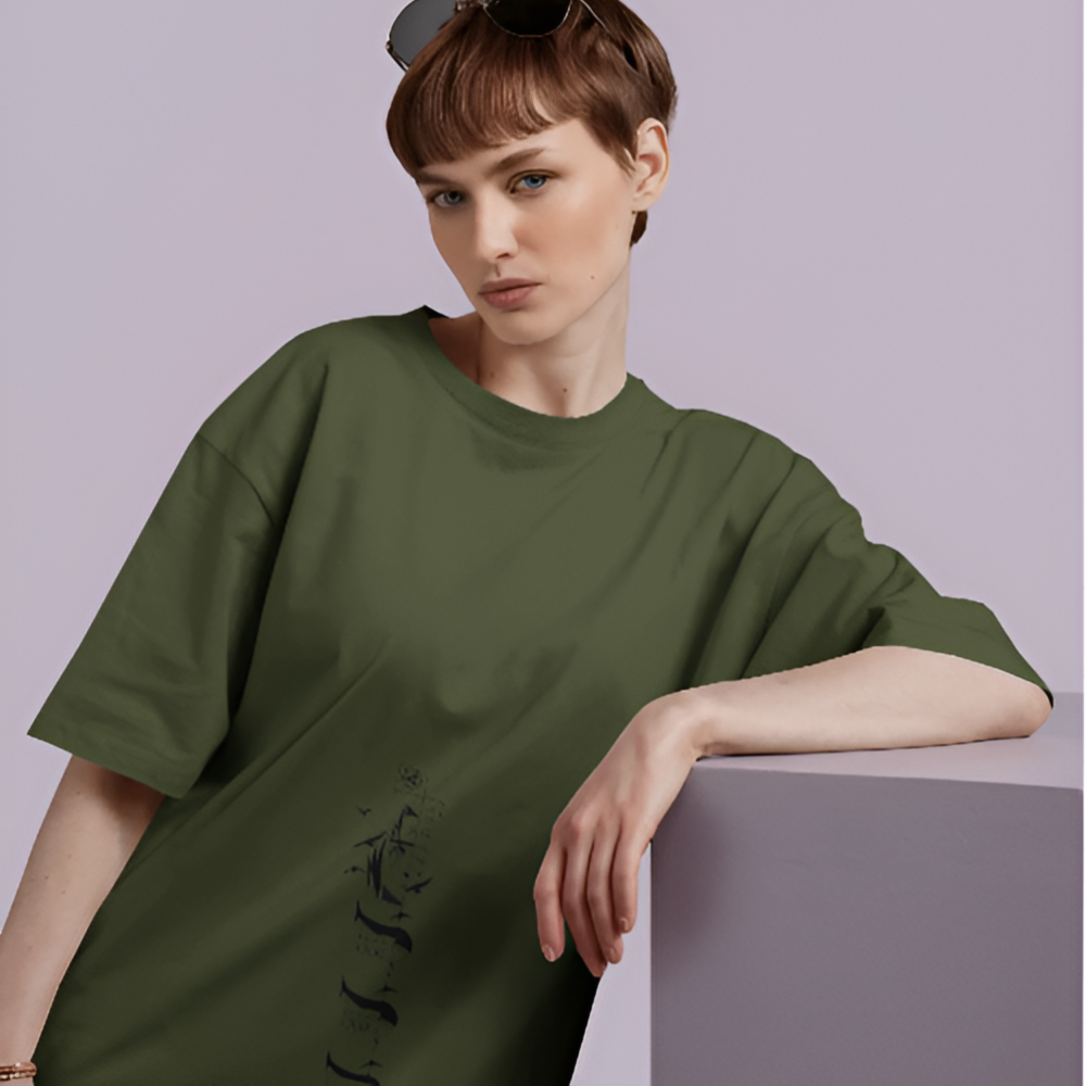A woman wearing an oversized olive green T-shirt with a subtle vertical graphic of a grim reaper surrounded by tombstones and crows, printed near the hem. She leans on a lavender block, wearing a calm yet edgy expression.