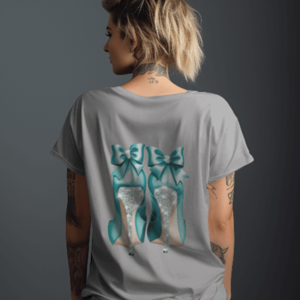 A woman facing away from the camera wearing a light grey T-shirt with a bold back graphic of glittery silver high heels topped with large teal bows. Her tattoos and soft undercut add an edgy contrast to the glam graphic.