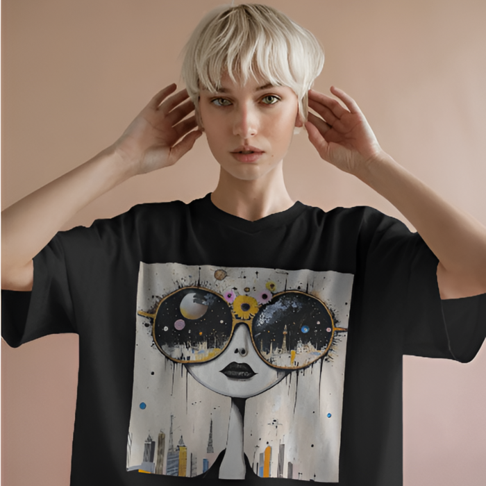 A woman with platinum blonde hair wearing a black oversized T-shirt featuring Cosmic City art print of a doll-like face with oversized round sunglasses that reflect galaxies and a glowing city skyline. The soft peach background contrasts with the bold shirt design.