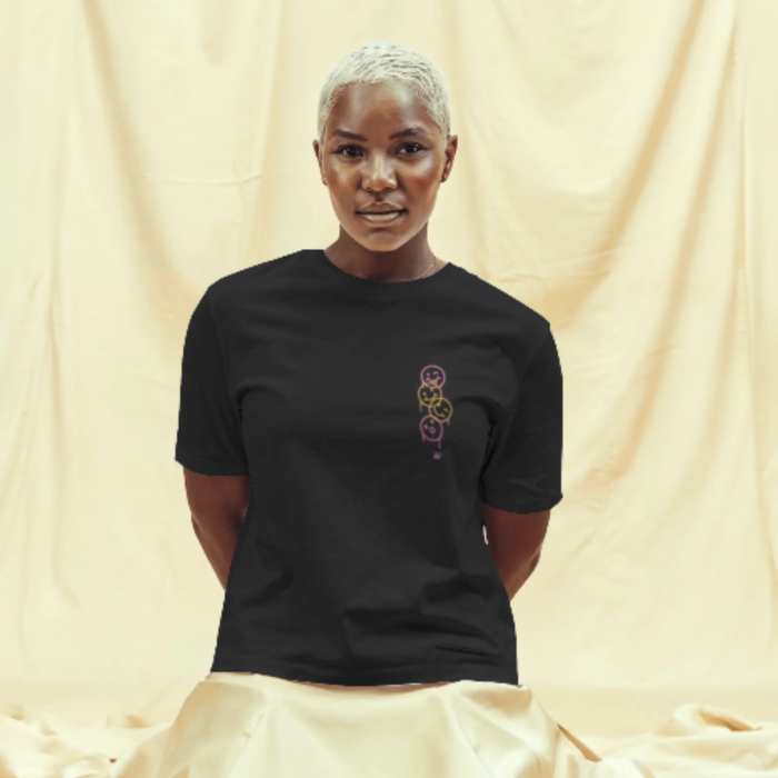 A woman wearing a black T-shirt with a small chest design of three minimalist faces stacked vertically in neon-style line art—one purple, one yellow, and one pink—each expressing a different emotion Live Your Best Life.