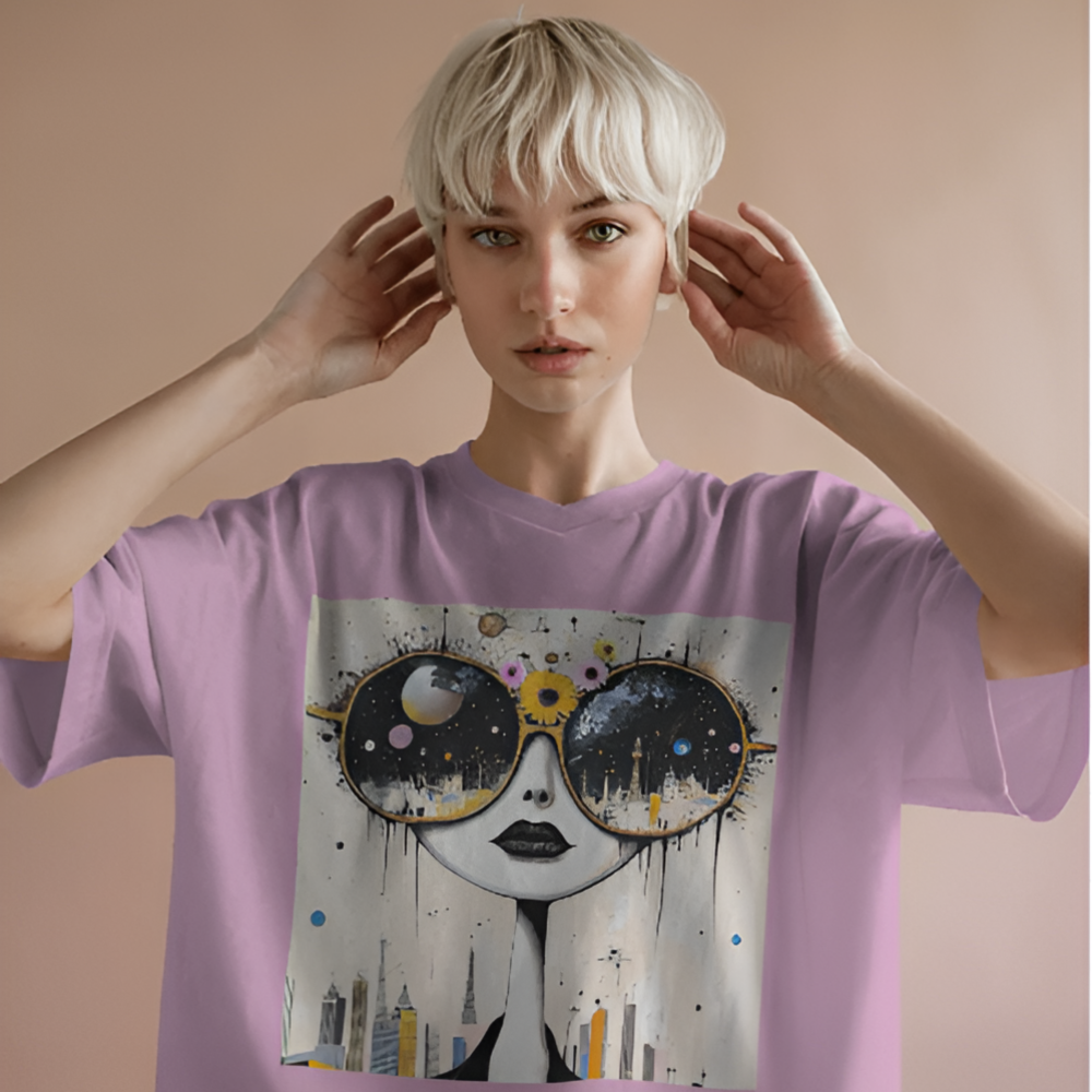 A woman wearing a lavender oversized T-shirt featuring a surreal art print of a doll-like face with oversized round sunglasses reflecting a cosmic night sky and a city skyline. The background is a soft peach-pink, and her pose is dreamy and relaxed.
