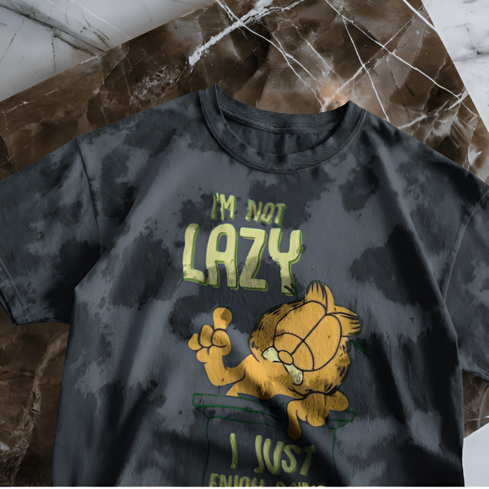 A black tie-dye T-shirt laid flat on a marble surface, featuring a retro-style illustration of Garfield with the quote “I’m not lazy, I just enjoy doing nothing” in green and yellow lettering. Garfield is resting on a ledge with his classic unimpressed expression.
