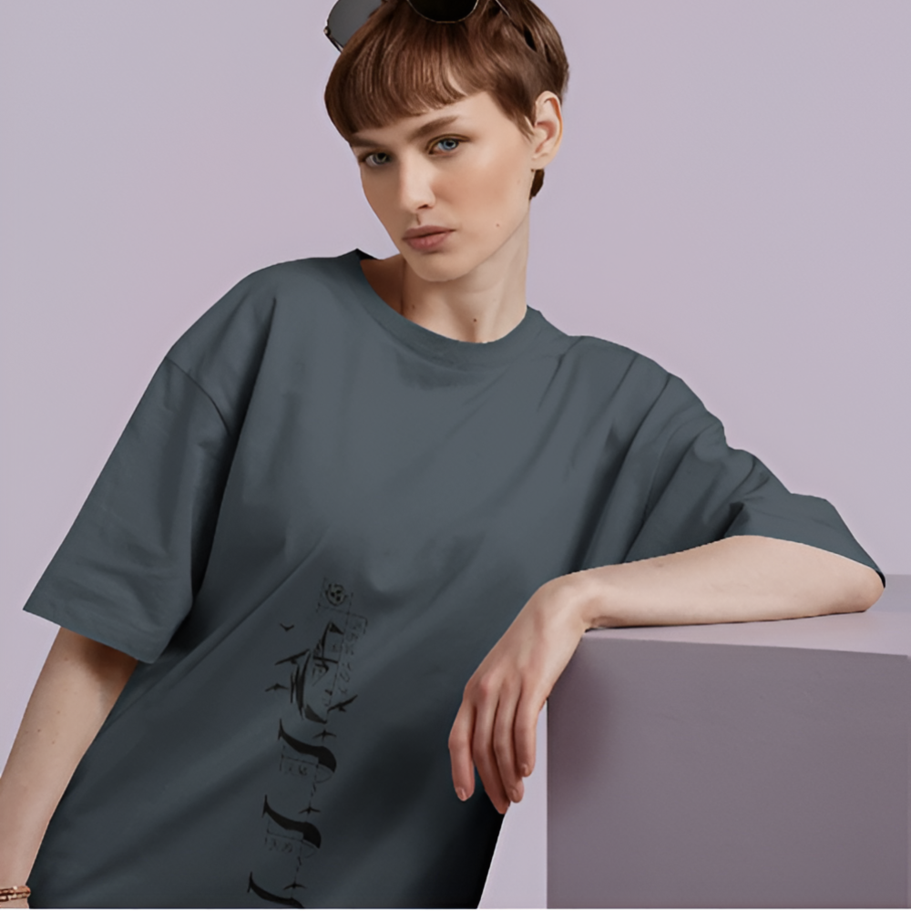 A woman wearing a loose-fit, dark charcoal T-shirt featuring a subtle vertical graphic of a grim reaper standing among tombstones, with flying birds and faded gothic elements. She leans stylishly against a lavender pedestal, exuding calm attitude.