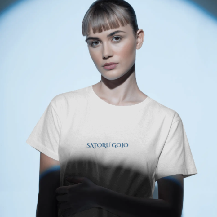 A woman wearing a white T-shirt with clean, minimal text in the center that reads "Satoru Gojo" in a sleek blue font. The spotlighted lighting gives the tee a cinematic, mysterious flair.
