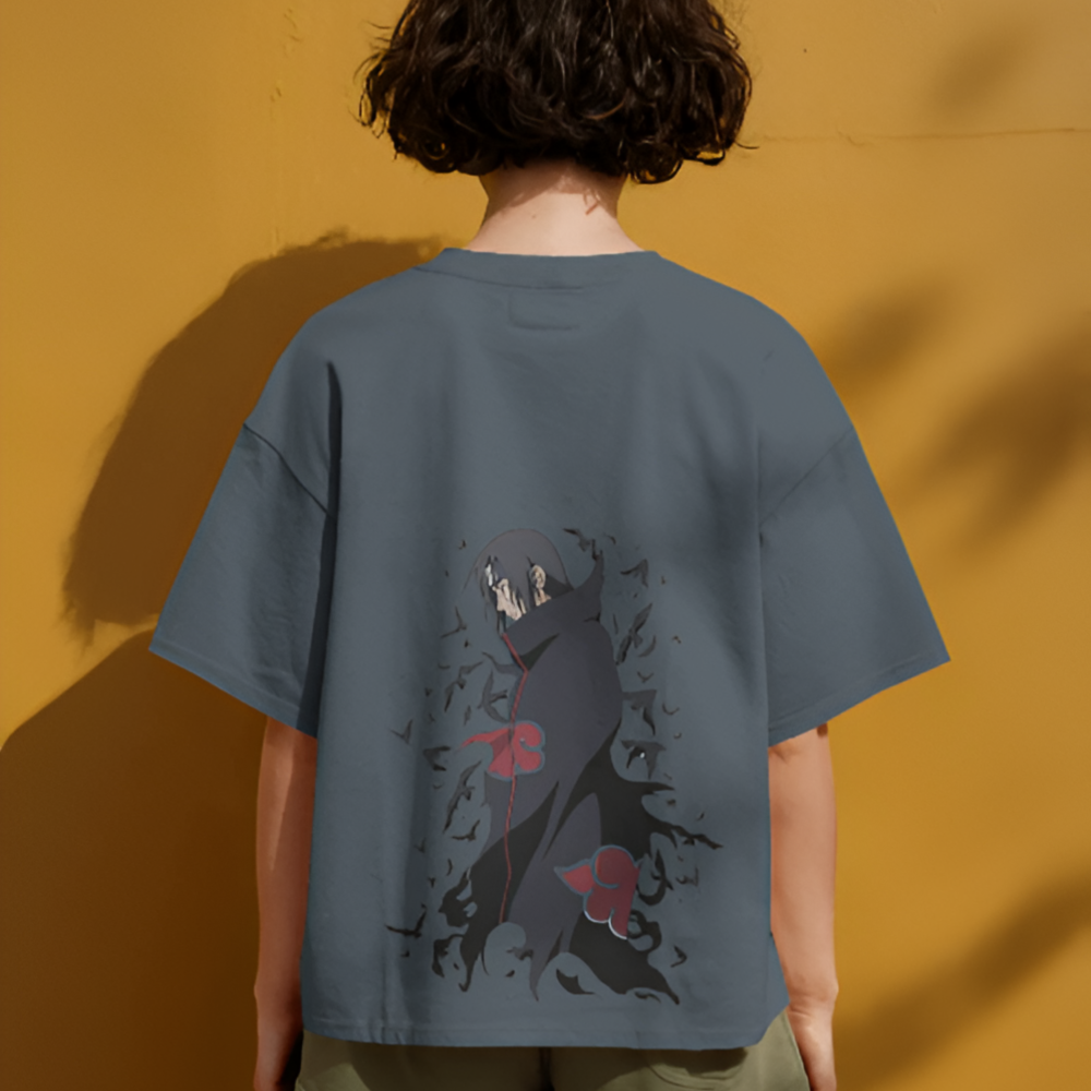 A person wearing a steely blue oversized T-shirt with a dramatic back graphic of Itachi Uchiha from Naruto. The character is cloaked in the Akatsuki robe, surrounded by crows and dark shadows, with red cloud symbols accenting the artwork. The mustard yellow wall creates a strong contrast.