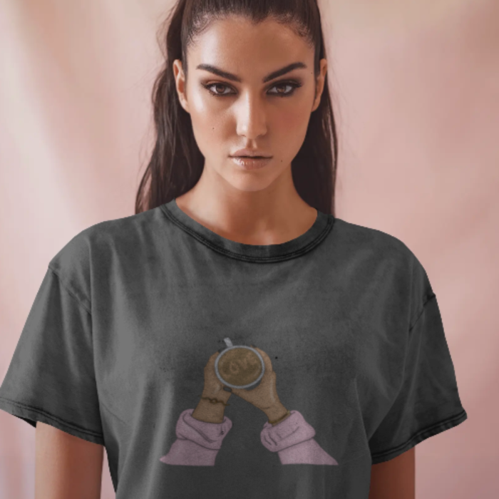 A woman wearing a black T-shirt featuring a centered illustration of two hands holding up a latte with “GRL” written in the foam. She stands against a soft pink background, radiating quiet strength.