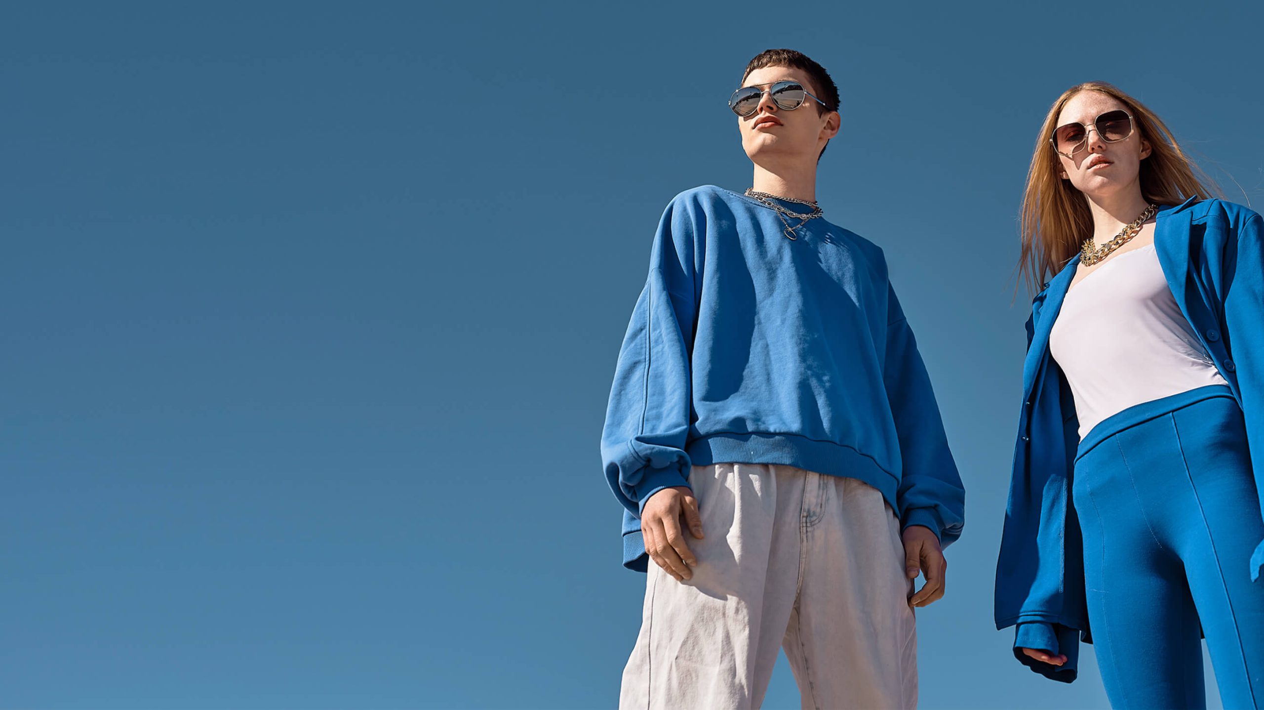 Two fashion models wearing blue-toned streetwear and sunglasses under a clear sky