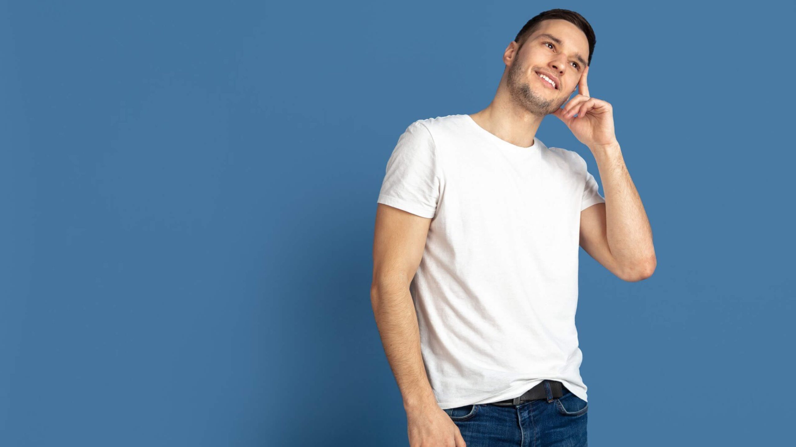 A Home smiling man in a white t-shirt and jeans, posing thoughtfully against a solid blue background.